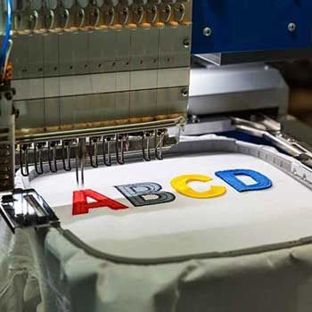 Máquina de bordado corporativo aplicando letras de colores sobre prenda blanca